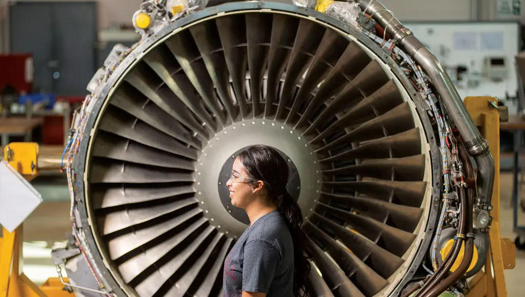 A student in safety glasses stands in profile at the center of a jet engine's fan blades