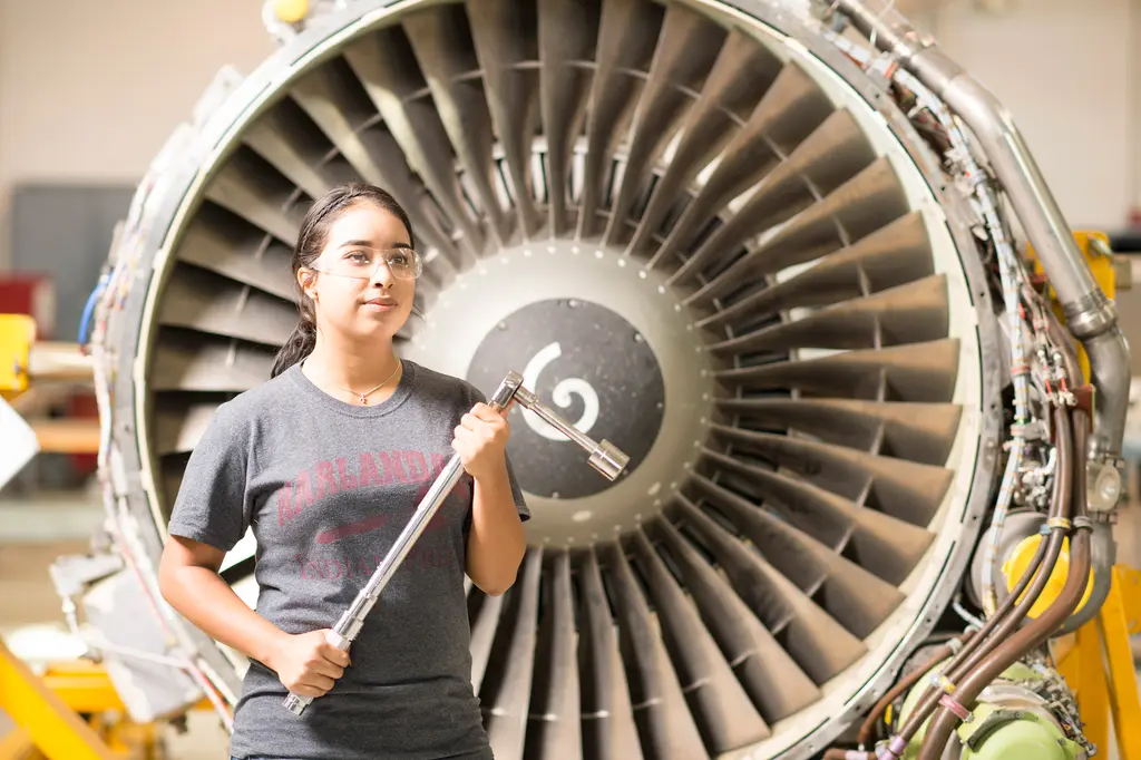 A student in safety glasses holds a large wrench in front of a jet engine in an aviation shop