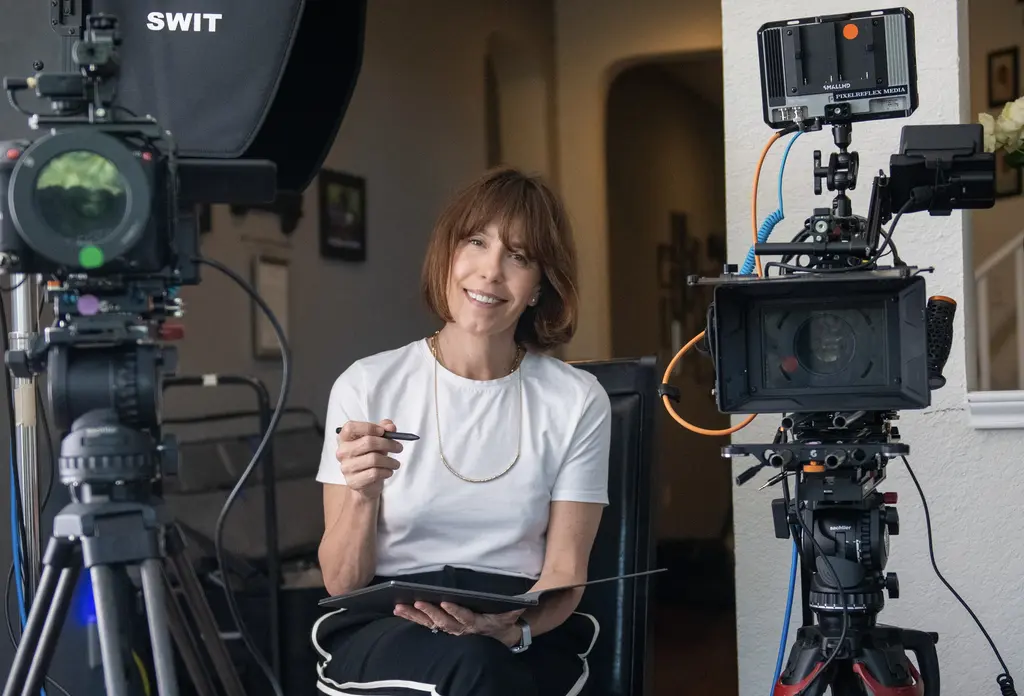 Steffi Ockenfels smiles while seated between two video cameras, holding a stylus, with a tablet on her lap.