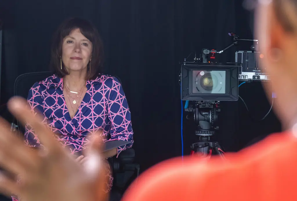 Steffi Ockenfels, con ropa estampada en azul y rosa, escucha sonriente junto a una cámara de cine; en primer plano, desenfocada, habla otra persona.