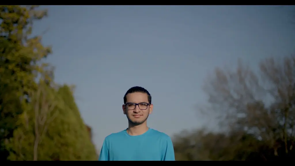 A Students of Service alum in glasses and a blue T-shirt stands outdoors against an evening sky.