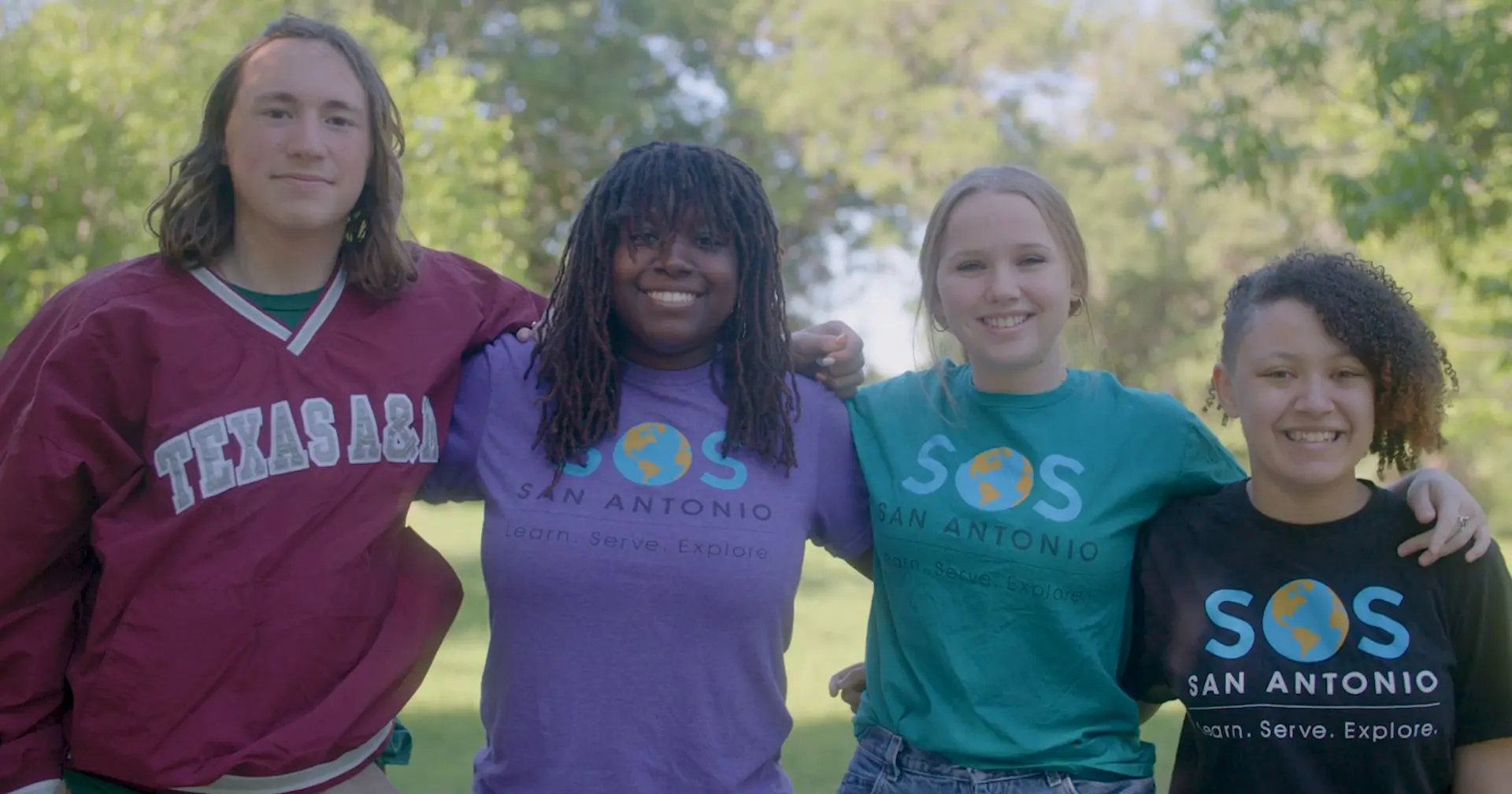 Students of Service member Trinity Williams, right, stands arm in arm with three other young people in SOS San Antonio shirts and a Texas A&M pullover under green trees.