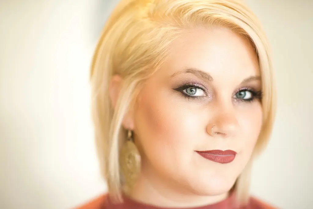 A woman with a side-swept blond bob, a nose ring and a large gold earring looks into the camera, in a rust-colored top against a soft cream background.