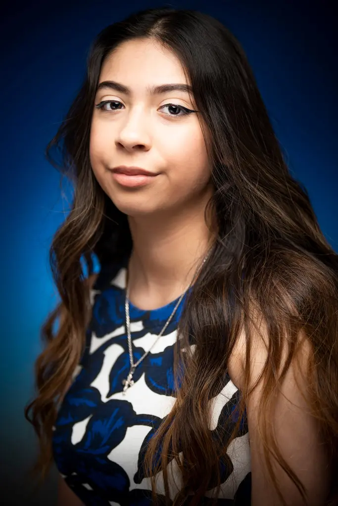 The same young woman in a blue-and-white floral dress and a silver cross necklace, turned slightly against the blue backdrop.