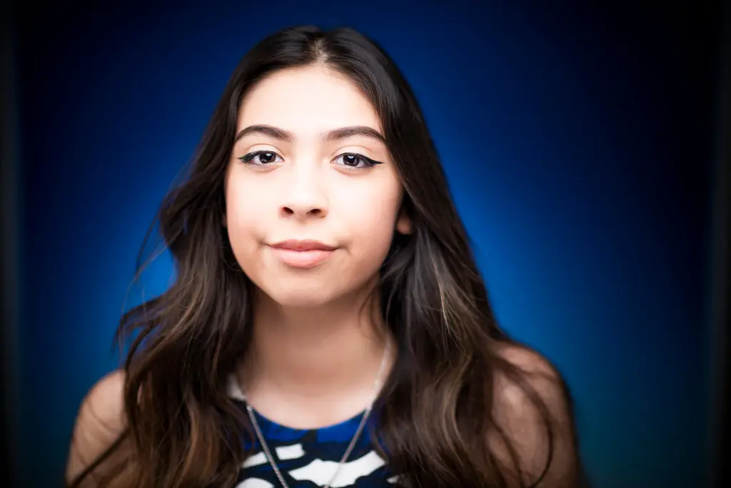 A young woman with long dark hair and winged eyeliner smiles softly against a deep blue backdrop.