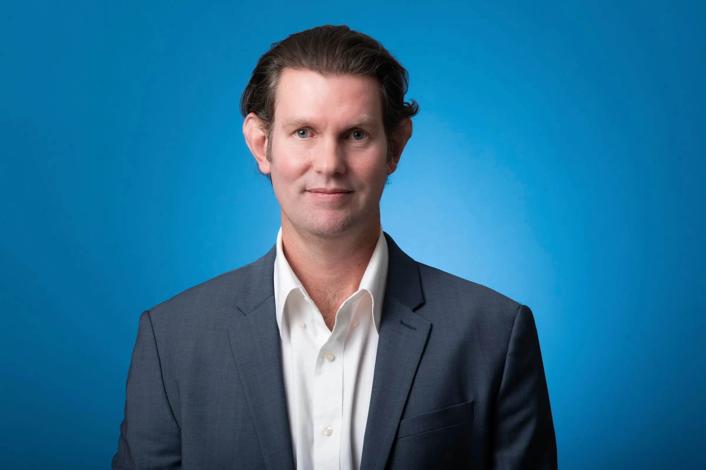 Sam Martens, president of Mission Solar Energy, in a charcoal suit and an open-collar white shirt, against a bright blue backdrop.
