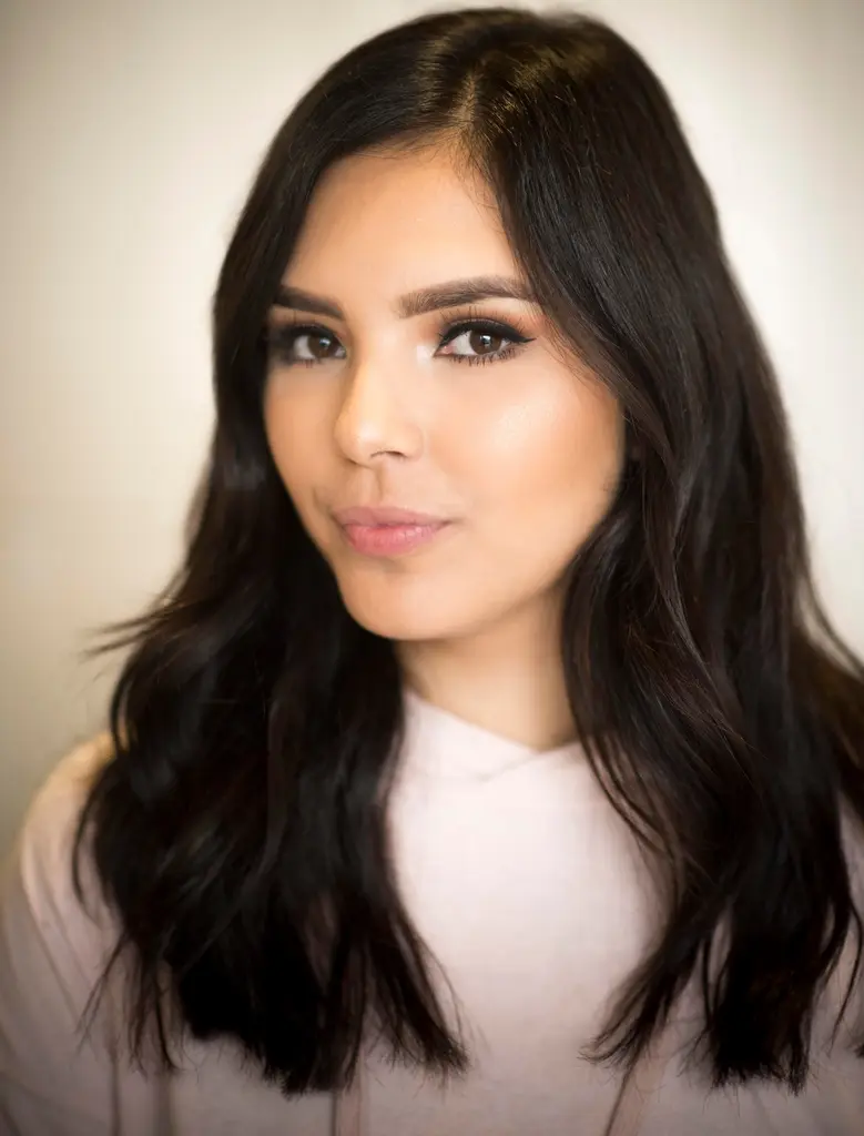 A woman with long, dark wavy hair and a slight smile, in a pale pink top, against a soft cream backdrop.