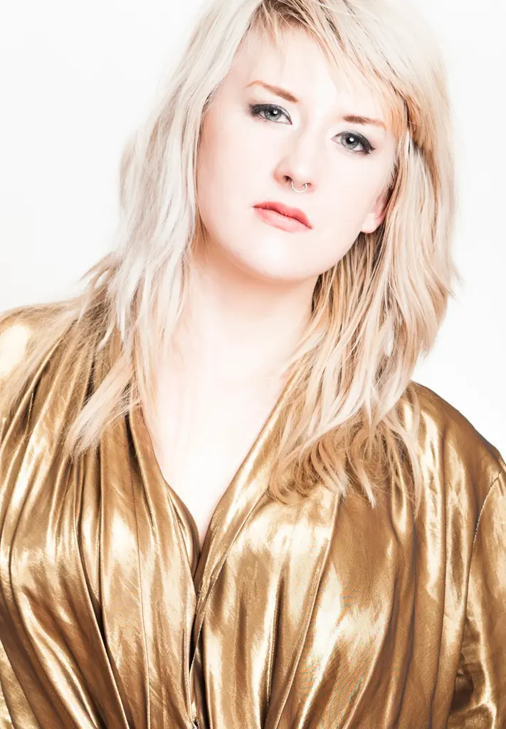 A woman with platinum blonde hair and a nose ring, in a gold lamé top, against a white backdrop.