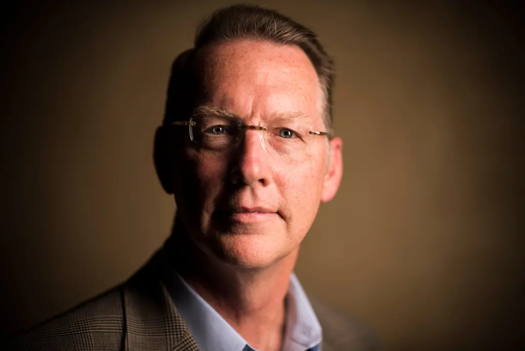 A man in rimless glasses and a plaid sport coat, lit with warm light against a dark brown backdrop.