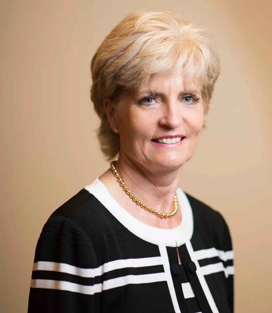 A smiling woman with short blonde hair, in a black-and-white cardigan and a gold bead necklace, against a warm beige backdrop.