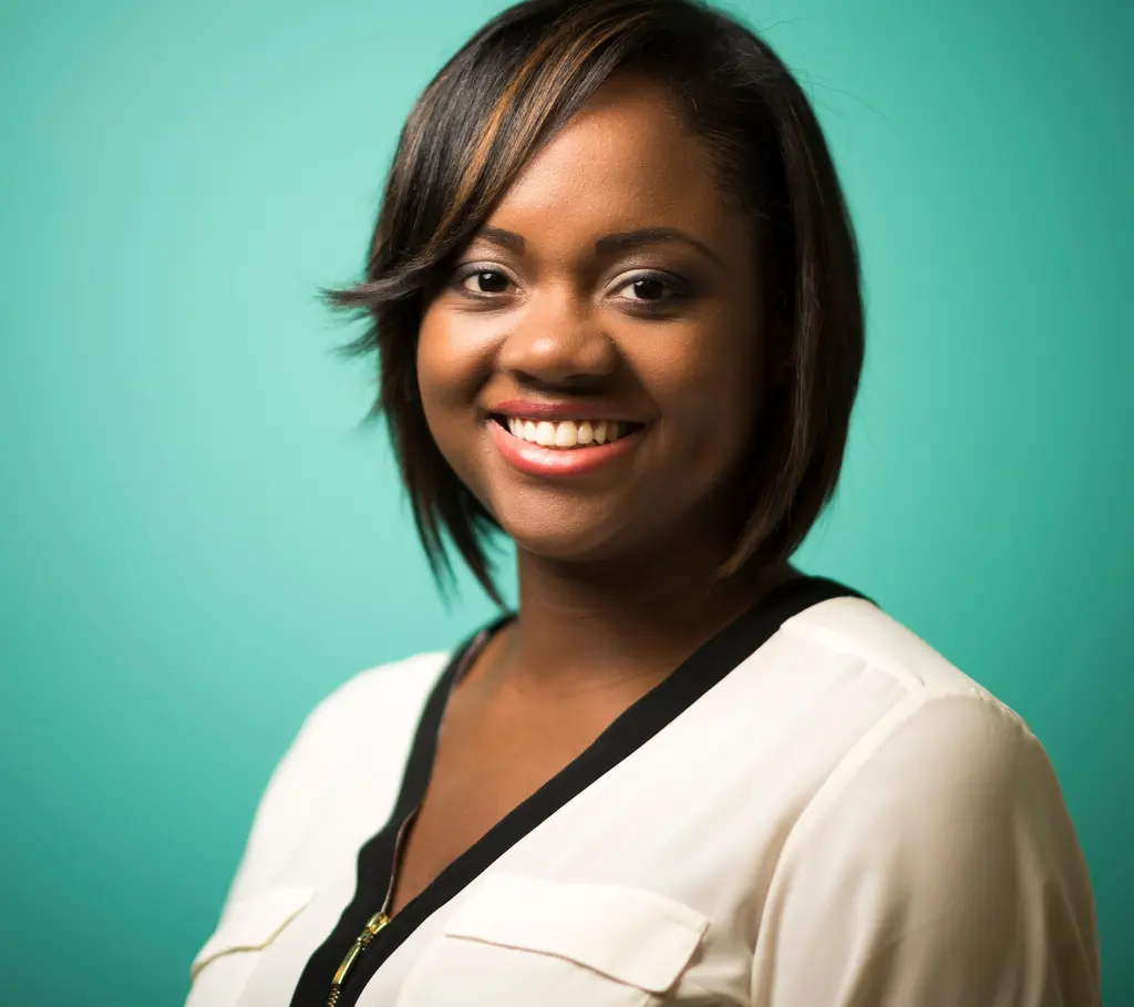 A smiling woman with a side-swept bob, in a cream blouse with black trim, against a teal backdrop.