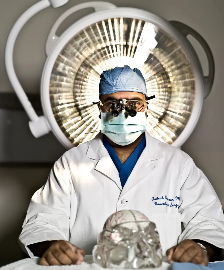 Neurosurgeon Sudesh Ebenezer in a surgical cap, mask and loupes sits under an operating light behind a glass skull