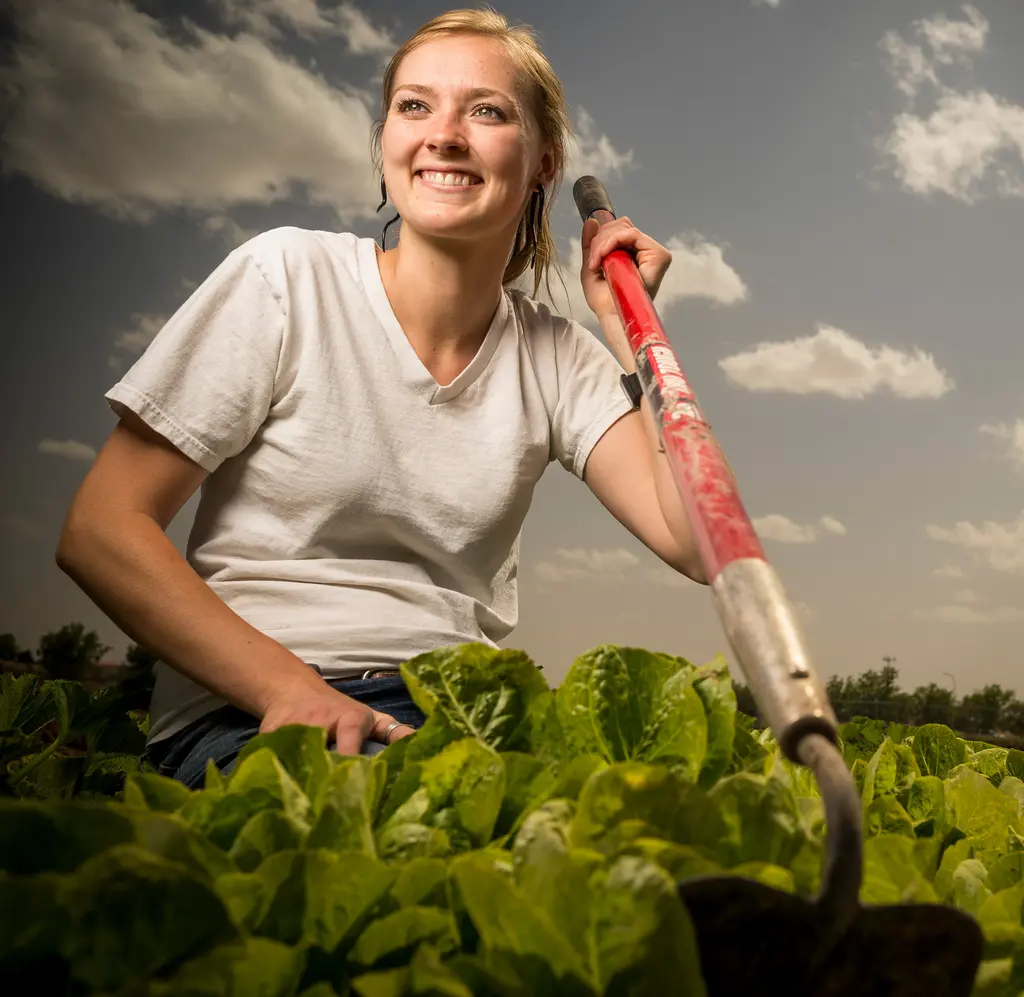 A smiling woman kneels with a hoe among rows of lettuce under a cloudy sky
