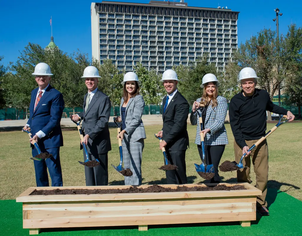 From left, Robert Thrailkill, David Zachry, Lori Houston, Ron Nirenberg, Brooke Thomas and Andres Andujar in white hard hats hold shovels of dirt behind a long wooden box of soil, with a tall hotel behind them.