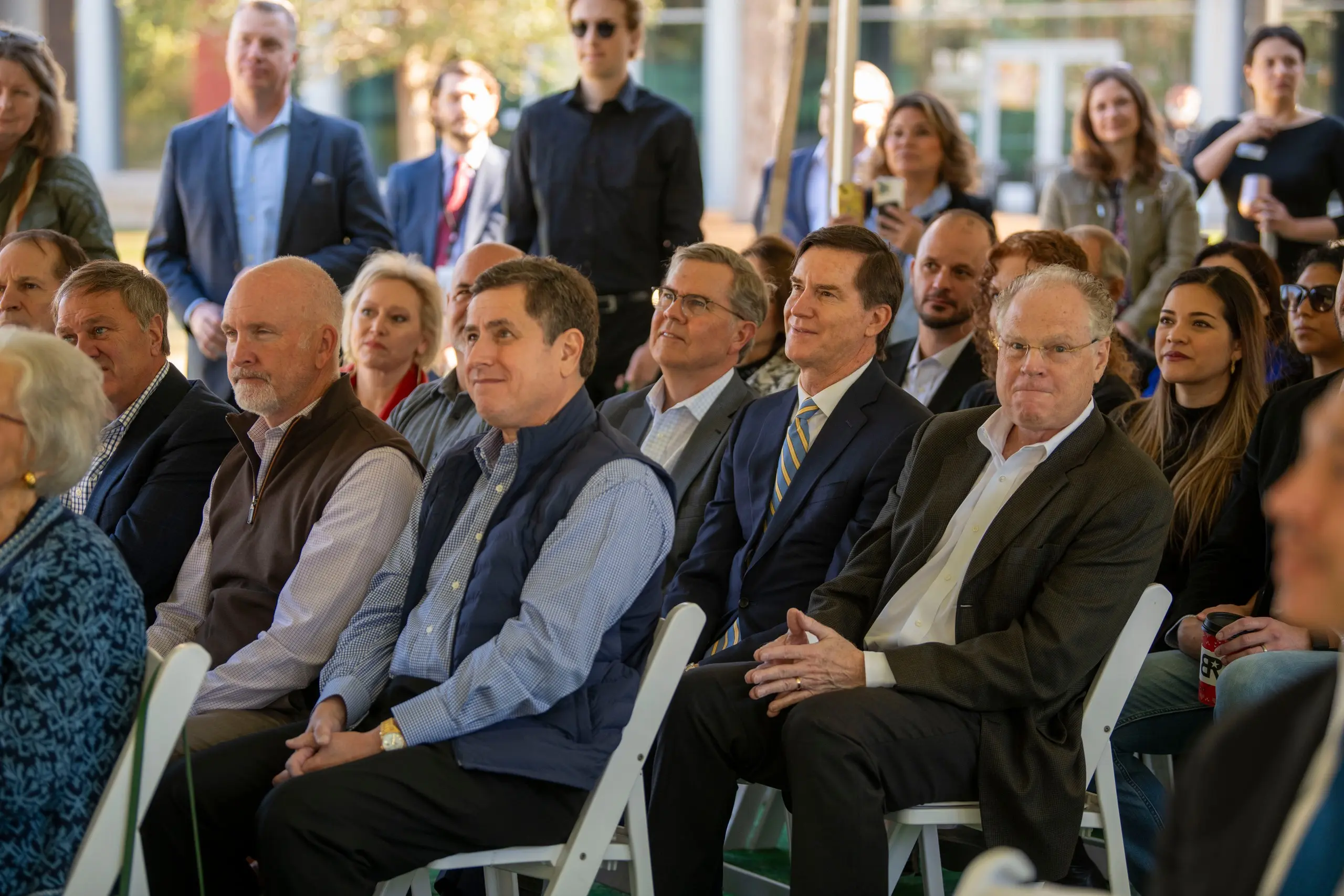 Rows of men and women in suits and vests sit on white folding chairs under a tent, looking toward the stage, with more guests standing behind them in the sun.
