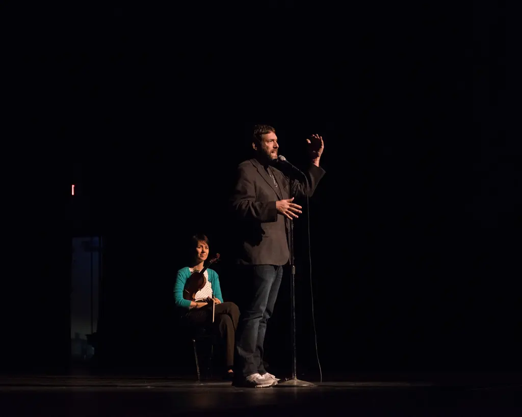 On a dark stage, a bearded man in a blazer, jeans and white sneakers gestures at a microphone while a woman in a teal cardigan sits holding a violin behind him to the left.