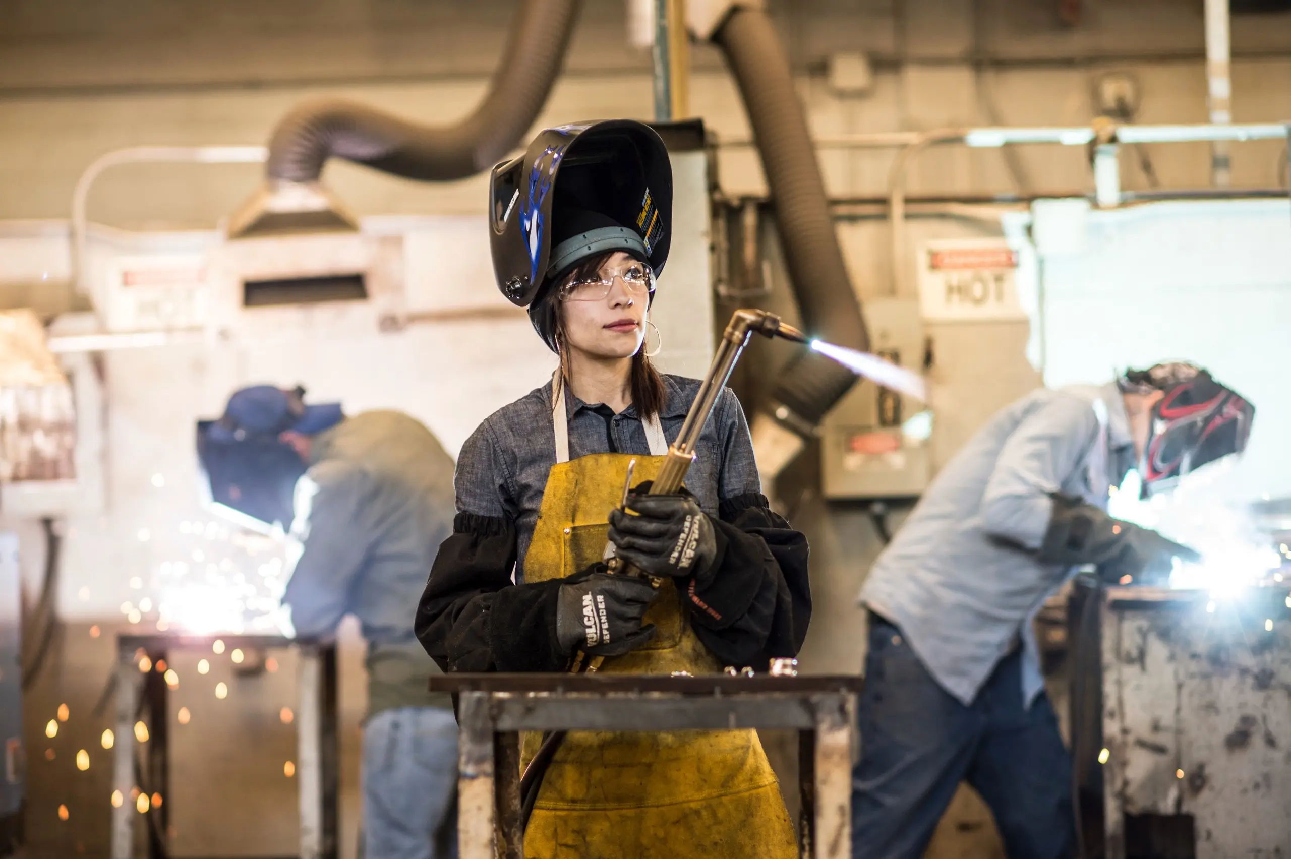 Woman in a flipped-up welding helmet and leather apron holds a torch as two welders work behind her amid sparks
