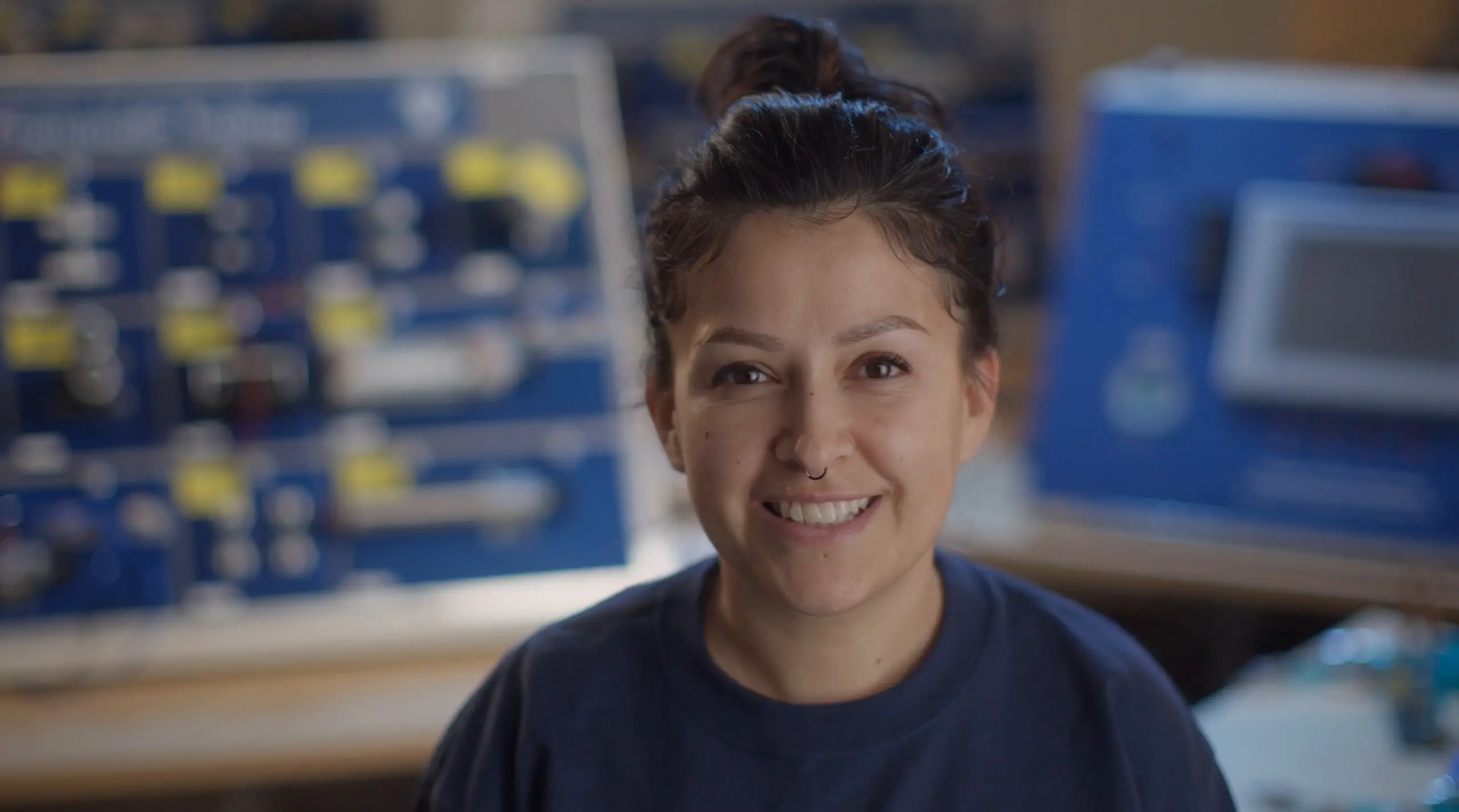 A smiling woman with her dark hair in a bun and a nose ring, wearing a navy T-shirt, in front of blurred blue training panels in a lab.