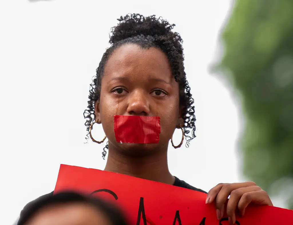 A young woman with red tape over her mouth cries as she holds a red sign during a campus demonstration.