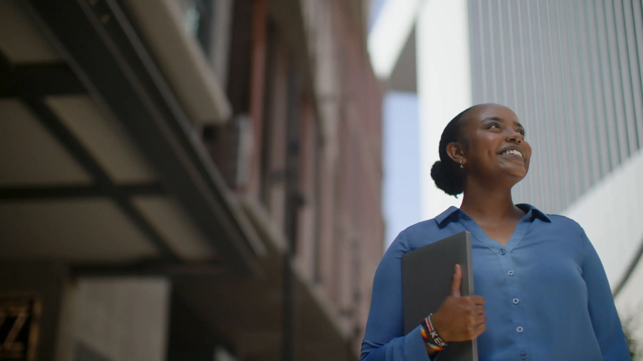 NPower Dallas alum Valentine Komen, in a blue shirt and holding a laptop, smiles as she looks up between city buildings.