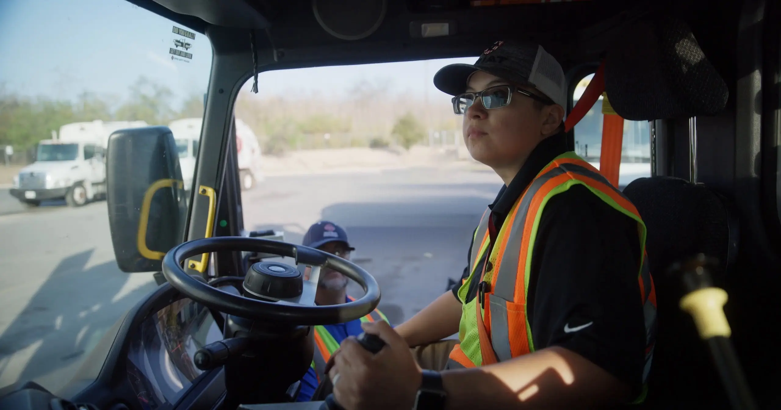 Valerie Perez, a City of San Antonio side loader operations lead in a safety vest, sits at the controls of a truck as a man in a blue cap rides along.