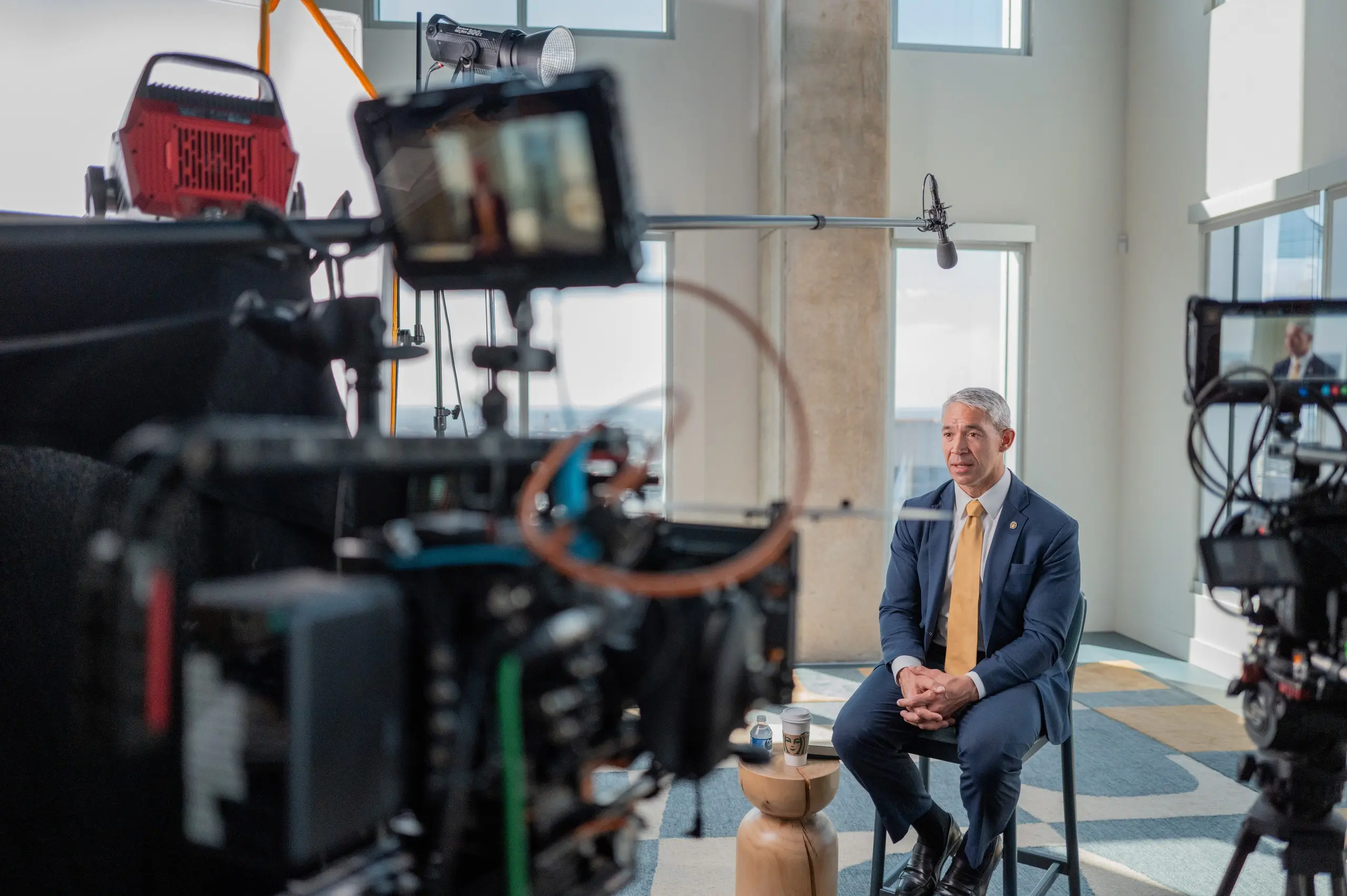 Former San Antonio Mayor Ron Nirenberg, in a navy suit and gold tie, sits for an interview by tall windows at 300 Main as cinema cameras and a boom mic record in the foreground.