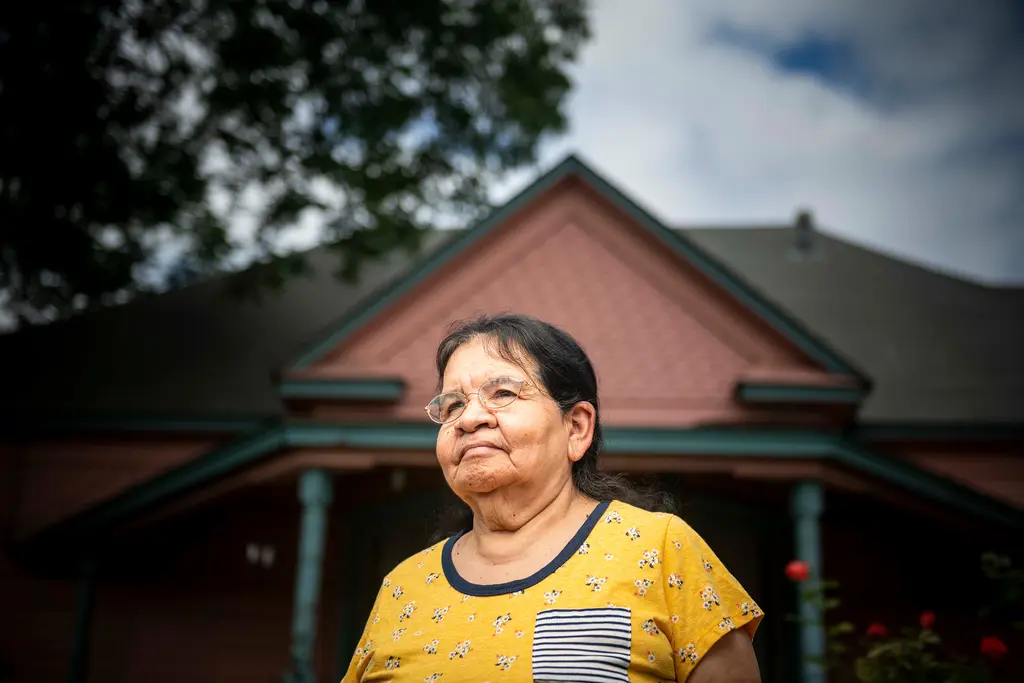 Virginia Guerra in a yellow top stands in front of her pink Victorian house, seen from a low angle