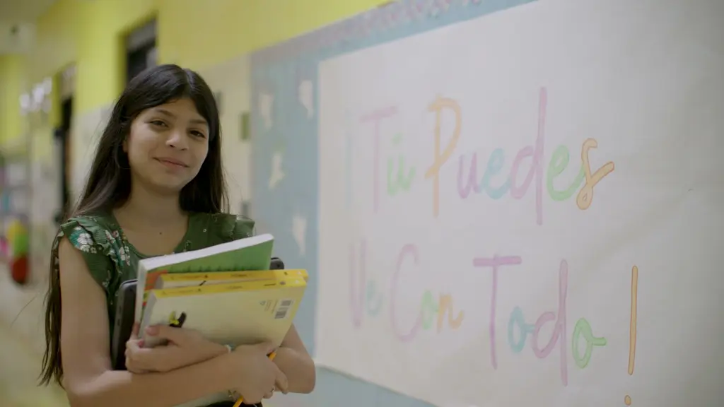 A girl in a green floral top holds a stack of books beside a hand-lettered sign that reads “¡Tú Puedes Ve Con Todo!”