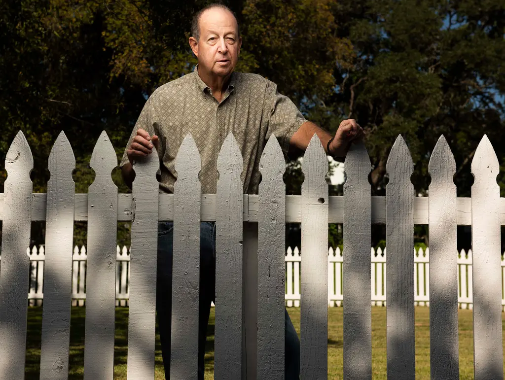 An older man rests his hands on the pointed tops of a white picket fence in dappled light.