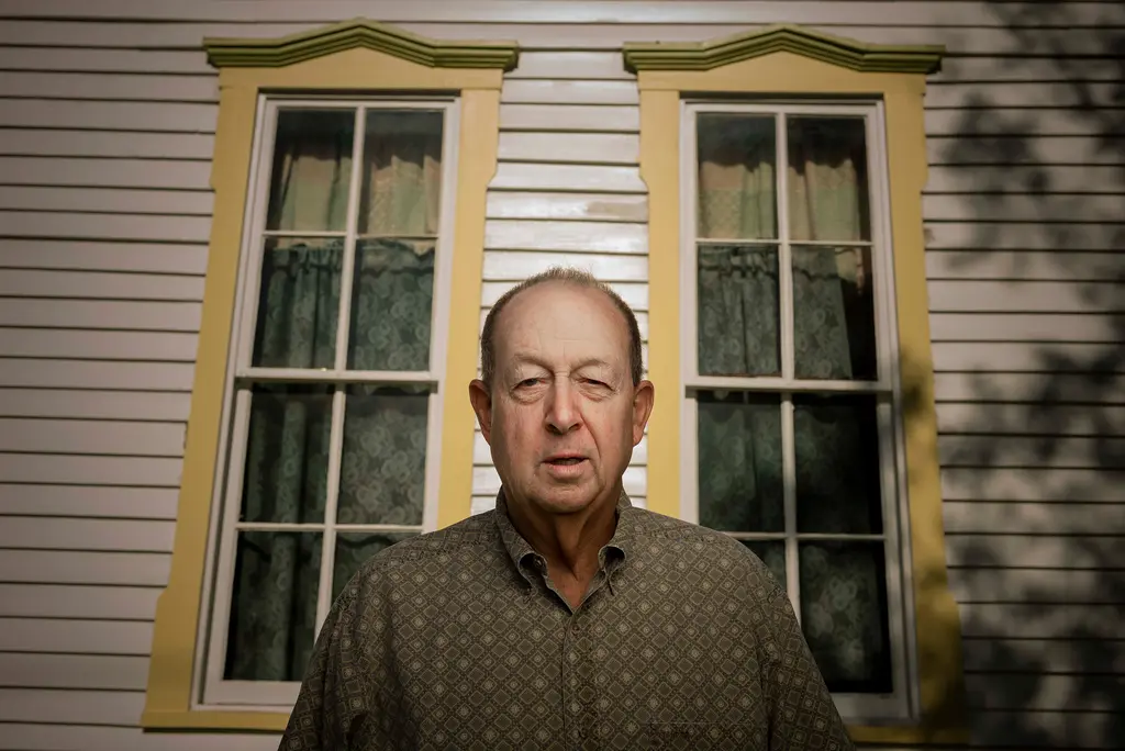 An older man in a patterned shirt stands in front of two tall windows with yellow trim on a white wooden house.
