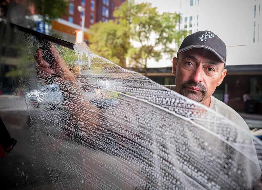 A window washer in a Spurs cap squeegees soapy water across a downtown storefront window