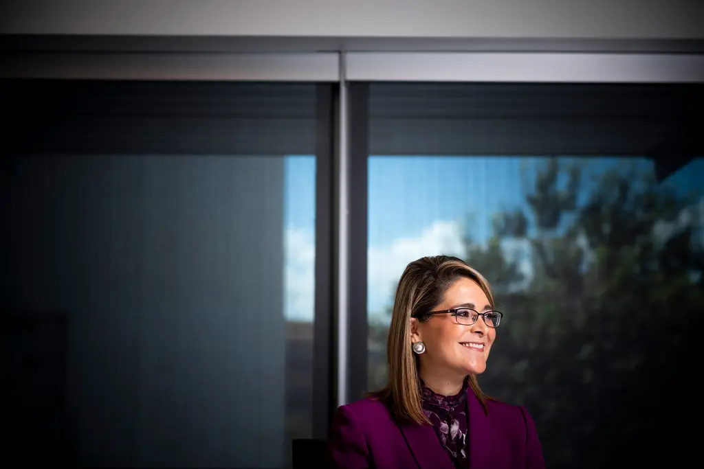 Xochy Hurtado, in a magenta suit and glasses, smiles as she looks out a tall window reflecting trees.