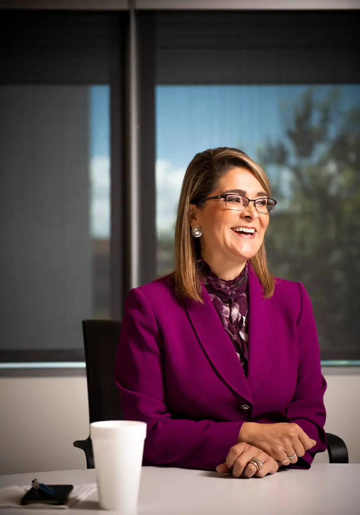 Xochy Hurtado in a magenta suit laughs while seated at a conference table by an office window