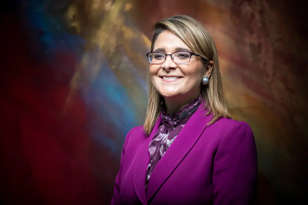 Xochy Hurtado in a magenta suit and glasses smiles in front of a painted backdrop