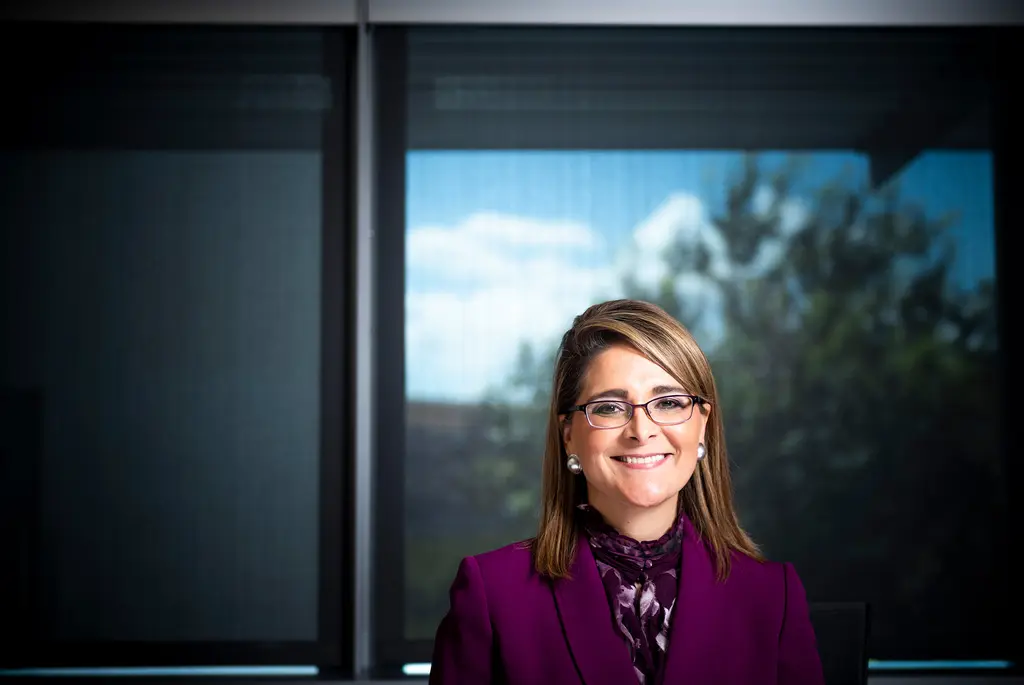 Xochy Hurtado, in a magenta suit and glasses, smiles in front of an office window with sky and trees outside.