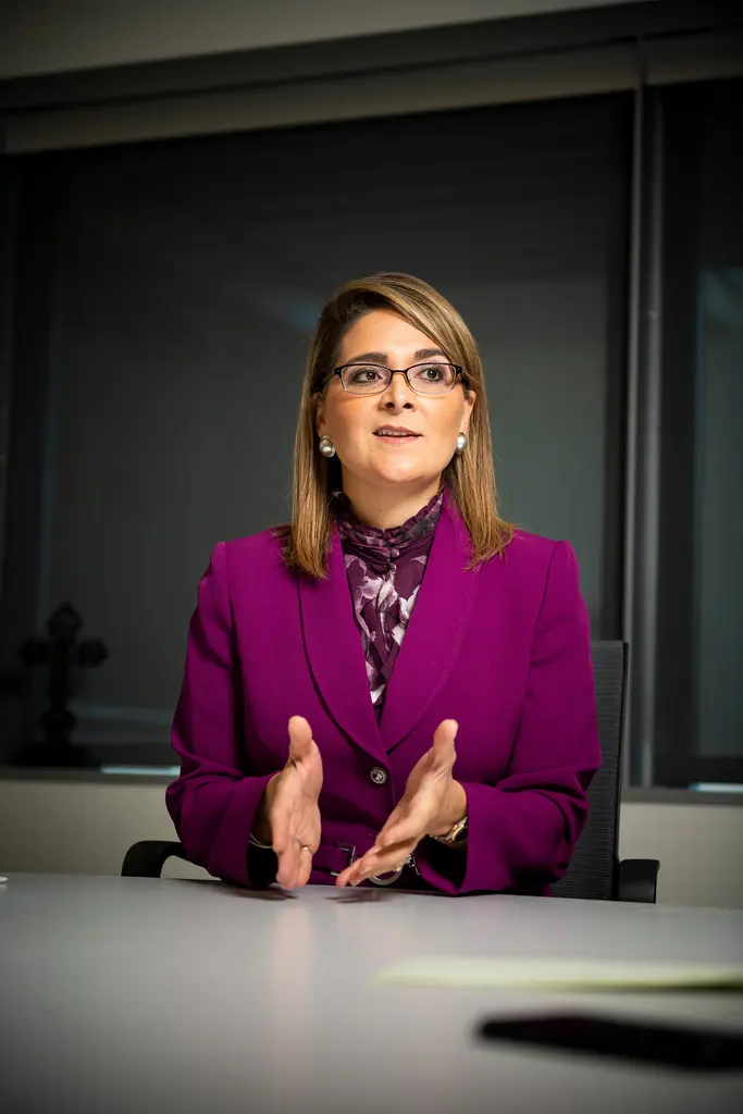 Xochy Hurtado, in a magenta suit, gestures with both hands as she talks at a white conference table.