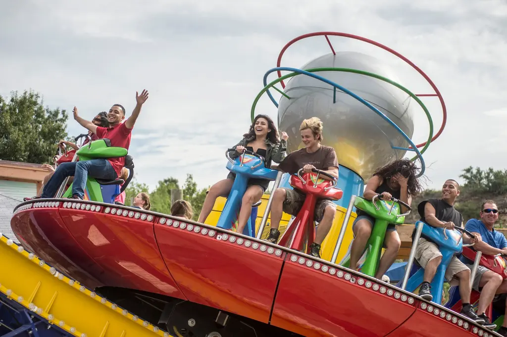 Riders in red, green and blue seats ride a tilted red disc under a silver globe ringed with colored hoops, one man raising both arms high and a woman beside him laughing.