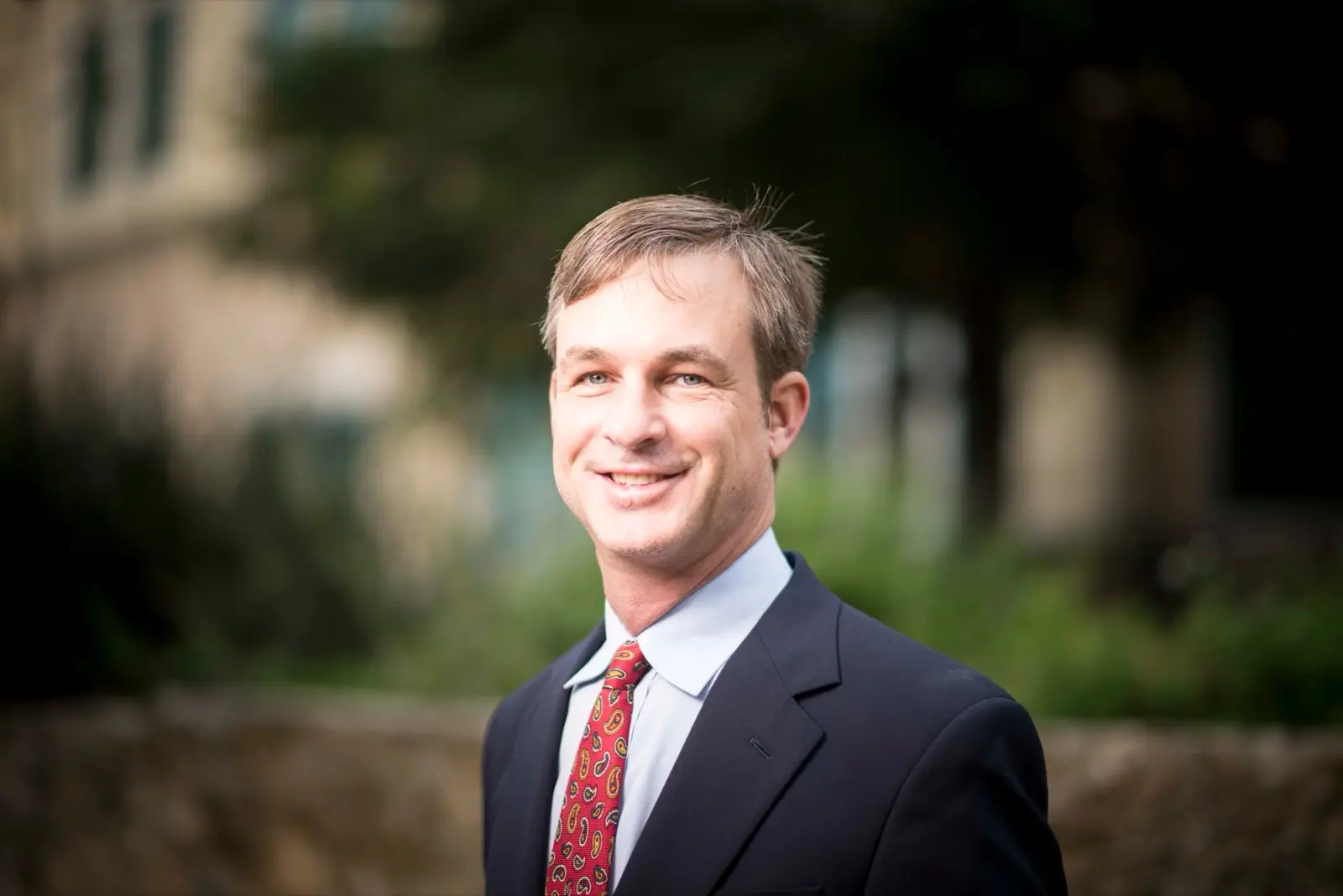 A smiling Zachry Construction employee in a navy suit and red patterned tie, photographed outdoors in front of trees and a building.